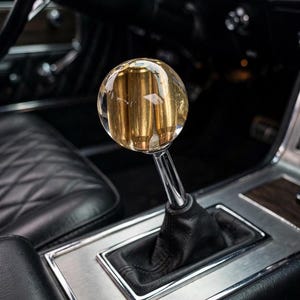 May include: A spherical gear shift knob featuring a clear resin exterior with gold-colored metallic bullet casings embedded inside. The knob is attached to a polished chrome metal shaft and a black leather boot, positioned within a vehicle interior.
