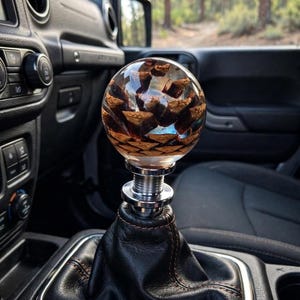 May include: A spherical gear shift knob featuring a natural brown pinecone encased in clear resin. The knob is mounted on a silver metal threaded base, which is attached to a black leather gear shift boot with visible brown stitching inside a vehicle.