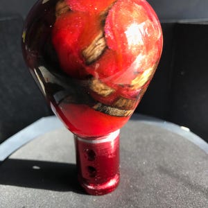 May include: A red and brown shift knob with a glossy finish. The knob has a rounded top with a wood-like pattern encased in a clear resin. The base is a solid red color with two small holes. Automotive accessory.