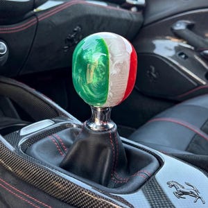 May include: A spherical gear shift knob featuring a glossy finish with vertical stripes in green, white, and red. The knob is mounted on a black leather shift boot with red stitching, set within a carbon fiber center console featuring a prancing horse logo.