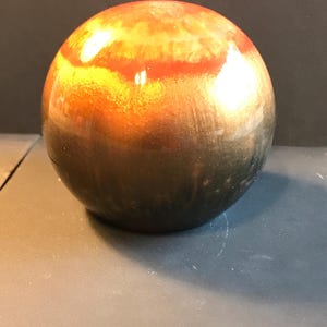 Puede incluir: Un objeto esférico pulido con un degradado de colores. La mitad superior pasa de un naranja vibrante a una banda roja oscura, mientras que la mitad inferior se desvanece en un marrón oscuro. La esfera es lisa y reflectante.
