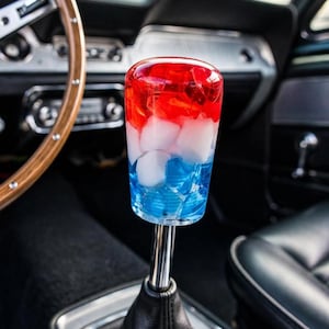 May include: A cylindrical gear shift knob made of clear resin featuring a layered design with a red top section, a white middle section containing embedded white fragments, and a blue bottom section, mounted on a chrome shaft with a black boot.