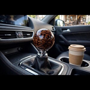 May include: A spherical gear shift knob made of clear resin containing dark brown roasted coffee beans. The knob is mounted on a black leather boot with visible stitching, positioned within a vehicle center console near a cup holder.