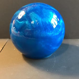 May include: A vibrant blue sphere with a glossy finish. The surface has a swirling, marbled effect with lighter blue and silver accents. The sphere is sitting on a gray surface.