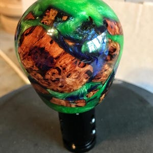 May include: A custom shift knob featuring a unique design. The top is a vibrant green, with sections of brown wood and blue accents. The knob is mounted on a black base, creating a striking contrast. This handcrafted piece adds a touch of artistry to any vehicle's interior.