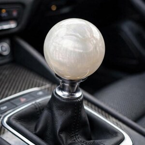 May include: A spherical gear shift knob with a pearlescent white finish, featuring a smooth, glossy surface. The knob is mounted on a silver-toned metal collar, which connects to a black leather shift boot within a vehicle's center console.