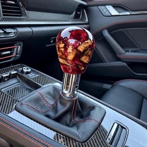 May include: A spherical gear shift knob featuring a blend of natural wood grain and deep red resin, mounted on a polished chrome shaft. The base is surrounded by a black leather boot with red stitching, set within a carbon fibre centre console.