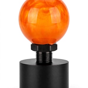 May include: A gear shift knob featuring a smooth, spherical orange resin top with swirling lighter orange patterns. The base is a solid, matte black cylindrical metal component with a threaded metal connector and a hexagonal nut attachment.
