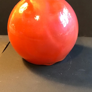 Puede incluir: Un objeto esférico de color rojo brillante con un acabado brillante. La superficie refleja la luz, creando reflejos. El objeto parece sólido y liso, con una base redondeada. El fondo oscuro acentúa el color y la forma.