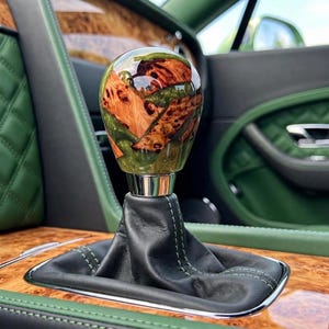 May include: A spherical gear shift knob featuring a polished resin finish with embedded wood grain patterns, mounted on a black leather boot with green stitching, situated within a vehicle interior with green quilted panels and wood trim.