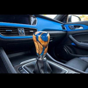 May include: A gear shift knob featuring a combination of natural light brown wood burl and vibrant blue resin. The knob is mounted on a black leather boot with blue stitching, positioned within a car interior with blue dashboard accents.
