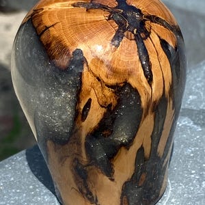 Puede incluir: Un objeto redondeado y bulboso elaborado con una combinación de madera natural de color marrón claro con vetas visibles y resina translúcida de color carbón oscuro. La madera presenta una grieta central oscura y formas orgánicas y fluidas.