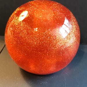 May include: A spherical, orange-red decorative object with a glittery, speckled surface. The sphere has a glossy finish and reflects light, creating bright highlights. The overall shape is a perfect sphere.