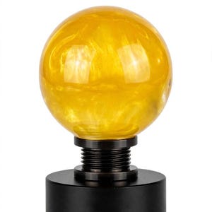 May include: A spherical yellow translucent glass orb mounted on a black cylindrical base. The orb features a swirling internal texture, while the base includes a ribbed neck section connecting the sphere to the solid black circular stand.