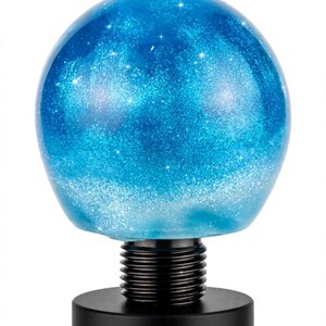 May include: A spherical gear shift knob featuring a translucent blue resin body filled with fine silver glitter that mimics a starry night sky. The sphere is attached to a black threaded metal base and a cylindrical black mounting component.