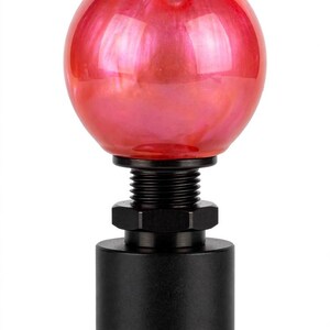 Può includere: Un pomello del cambio caratterizzato da una sfera in vetro rosso liscio con finitura perlata. La base è realizzata in metallo nero opaco, con un collo cilindrico filettato e un dado esagonale per un'installazione e regolazione sicura.