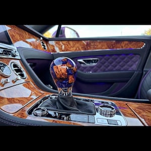 May include: A spherical gear shift knob featuring a polished finish with a swirling pattern of deep purple resin and natural brown wood burl. The knob is mounted on a black leather boot within a vehicle interior with wood grain trim and purple accents.