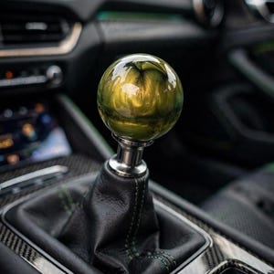 May include: A spherical gear shift knob with a glossy, swirling olive green and gold finish, mounted on a metallic silver base. The knob is attached to a black leather shift boot featuring visible green stitching, situated in a car interior.