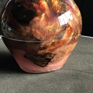 May include: A decorative, spherical object with a unique, swirling design. The top portion features a blend of gold, brown, and red hues, while the base is a soft pink. The glossy surface reflects light, enhancing the visual appeal.