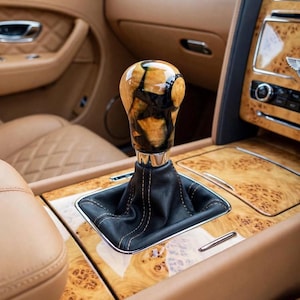 May include: A gear shift knob featuring a polished burl wood and black resin design, mounted on a black leather boot with visible stitching. The knob is positioned within a vehicle interior that includes tan leather upholstery and burl wood trim.