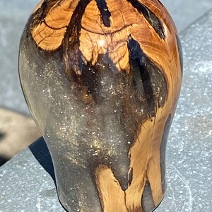 May include: A rounded gear shift knob crafted from a combination of natural light brown wood with visible grain patterns and translucent dark gray resin infused with fine gold-colored glitter particles throughout the lower section.