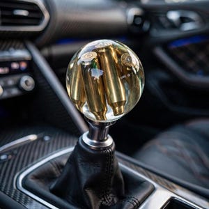 May include: A spherical gear shift knob made of clear resin, featuring several brass-coloured metallic bullet casings embedded inside. The knob is mounted on a silver-toned metal base attached to a black leather gear shift boot inside a vehicle.