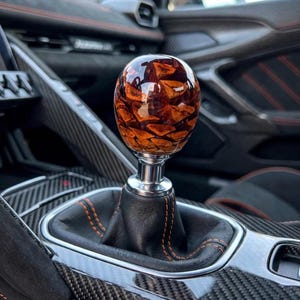 May include: A spherical gear shift knob featuring a polished, glossy resin finish with embedded brown and orange pinecone scales. The knob is mounted on a silver-toned metal base, attached to a black leather shift boot with orange stitching.
