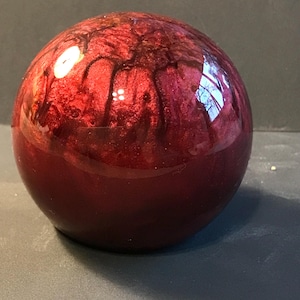 May include: A glossy, spherical object with a deep red hue, featuring intricate dark red marbling patterns. The surface reflects light, creating highlights. The object appears to be made of a solid, polished material.