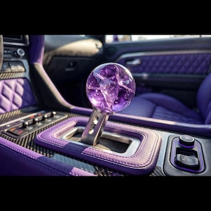 May include: A spherical purple gear shift knob with a cracked glass texture mounted on a metallic silver stem. The gear shifter is positioned within a vehicle interior featuring purple quilted leather upholstery and carbon fiber trim accents.