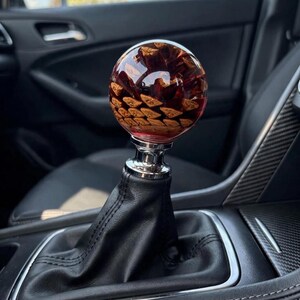May include: A spherical gear shift knob featuring a natural brown pinecone encased in clear, glossy resin. The knob is mounted on a polished silver-tone metal base, which is attached to a black leather gear shift boot inside a vehicle interior.