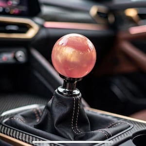 May include: A spherical gear shift knob with a glossy, translucent pink and gold marbled finish, mounted on a black leather shift boot with visible stitching, positioned within the center console of a vehicle interior.