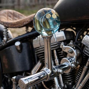 Op de afbeelding: Een verchroomde motorversnellingspook met een bolvormige knop van heldere hars. De knop bevat ingebedde messing kogelhulzen en een stuk zilverkleurig prikkeldraad. De pookarm is van gepolijst metaal met een cilindrische handgreep.