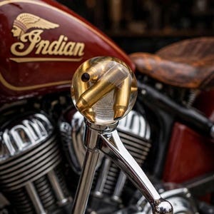 May include: A spherical clear resin gear shift knob containing brass-colored bullet casings, mounted on a polished chrome metal lever. The knob is positioned in front of a dark red motorcycle fuel tank with the gold Indian logo and engine parts.