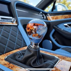 May include: A spherical gear shift knob featuring a swirling blend of blue and orange resin with wood grain accents, mounted on a black leather boot with blue stitching, situated within a car interior with blue quilted leather and wood trim.