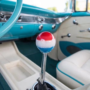May include: A spherical gear shift knob featuring horizontal bands of red, white, and blue, mounted on a polished chrome metal shaft. The knob is positioned within the interior of a vintage vehicle with a turquoise and white dashboard and seating.