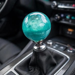May include: A spherical gear shift knob made of translucent teal resin with a swirling internal pattern, mounted on a metallic silver base atop a black leather gear shift boot inside a vehicle interior with dark trim and dashboard controls.