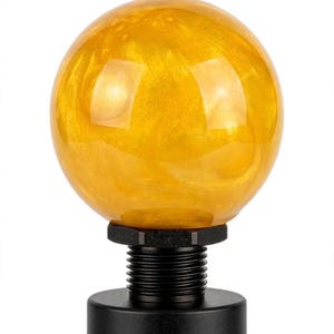 May include: A spherical, translucent amber-colored resin finial with a swirling, pearlescent texture. The orb is mounted on a black, cylindrical metal base featuring a threaded neck and a flat, circular collar at the point of attachment.