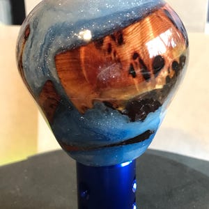 Può includere: Un pomello del cambio personalizzato con un design blu e venature del legno. La parte superiore è un mix di resina blu e legno, con un effetto glitterato. La base è in metallo blu brillante con fori.