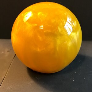 Puede incluir: Un objeto esférico brillante de color amarillo dorado. La superficie refleja la luz, creando reflejos y un aspecto brillante. La esfera parece estar hecha de un material sólido, posiblemente vidrio o resina.