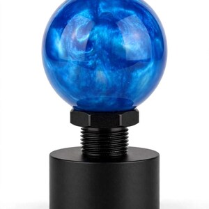 May include: A spherical gear shift knob featuring a swirling blue and white marbled pattern. The top sphere is attached to a black metal cylindrical base with a threaded neck and a hexagonal nut, providing a secure mounting mechanism for installation.
