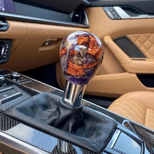 May include: A spherical gear shift knob featuring a polished finish with embedded pieces of natural brown wood grain and translucent purple resin. The knob is mounted on a metallic silver shaft with a black leather boot base in a vehicle interior.