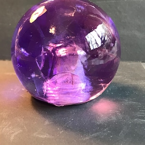 Puede incluir: Un objeto esférico translúcido de color púrpura intenso. El orbe de vidrio refleja la luz, creando reflejos y sombras. La parte inferior de la esfera tiene un tono más claro, rosado. El objeto parece estar hecho de vidrio o un material similar.