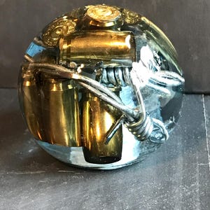 May include: A clear, spherical paperweight with embedded brass bullet casings and barbed wire. The metallic elements are encased in a transparent resin, creating a unique, industrial-themed decorative object.