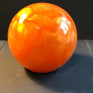 May include: A glossy, orange sphere with a reflective surface. The spherical object has a smooth texture and appears to be made of a solid material. The color is a vibrant, warm orange with hints of yellow.