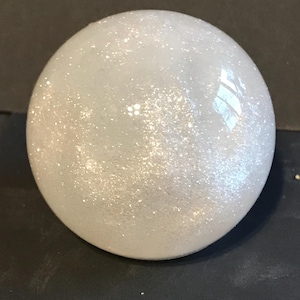 May include: A round, white decorative sphere with a glittery, iridescent finish. The sphere is smooth and reflective, catching the light. The object is sitting on a dark surface.