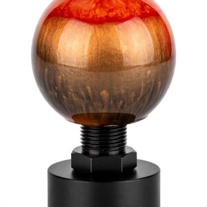 May include: A spherical gear shift knob featuring a glossy finish with a gradient of vibrant red at the top transitioning into a metallic bronze and dark brown base. The knob is attached to a black threaded metal post and a cylindrical black base.