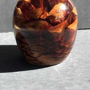 以下が含まれることがあります： 乾燥した赤茶色の松ぼっくりの鱗で満たされた、透明で丸みを帯びた装飾品。光沢のある仕上がりで、灰色の表面に置かれています。透明な外側を通して松ぼっくりの鱗が見えます。