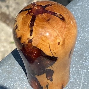 Puede incluir: Un objeto pulido en forma de cúpula hecho de madera de color marrón claro con una incrustación de resina de color marrón rojizo oscuro. La veta de la madera es claramente visible, con grietas naturales y patrones rellenos de resina oscura.