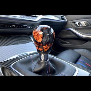 Può includere: Un pomello del cambio con una parte superiore arrotondata e lucida, caratterizzato da un motivo vorticoso di venature del legno arancioni e resina nera con granelli di glitter argento. È fissato a una cuffia in pelle nera con cuciture blu e rosse.