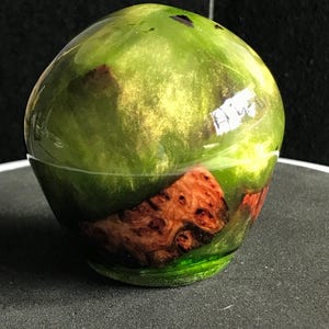 Puede incluir: Un cuenco decorativo hecho a mano, de forma redondeada y brillante. El cuenco es predominantemente verde con reflejos dorados y una sección de veta de madera natural. La base es de color verde oscuro, creando un contraste. El cuenco se utiliza probablemente con fines decorativos.